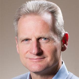 Greg Farrar wearing a suit and tie smiling and looking at the camera